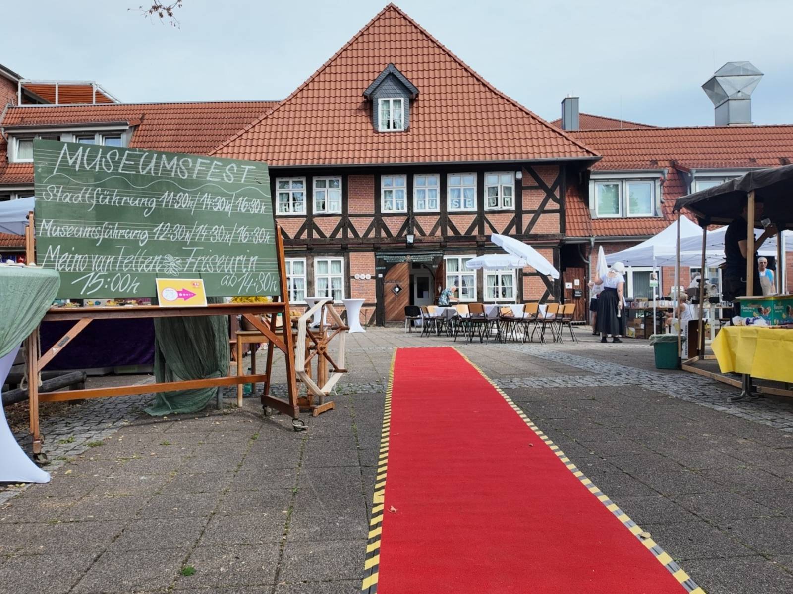 In Richtung des Gebäudes des Heimatmuseums liegt ein roter Teppich. Neben dem roten Teppich steht eine alte Schultafel, auf der die Zeiten anstehender Führungen stehen. Vor dem Museumsgebäude stehen zahlreiche Tische, Stühle und Sonnenschirme.