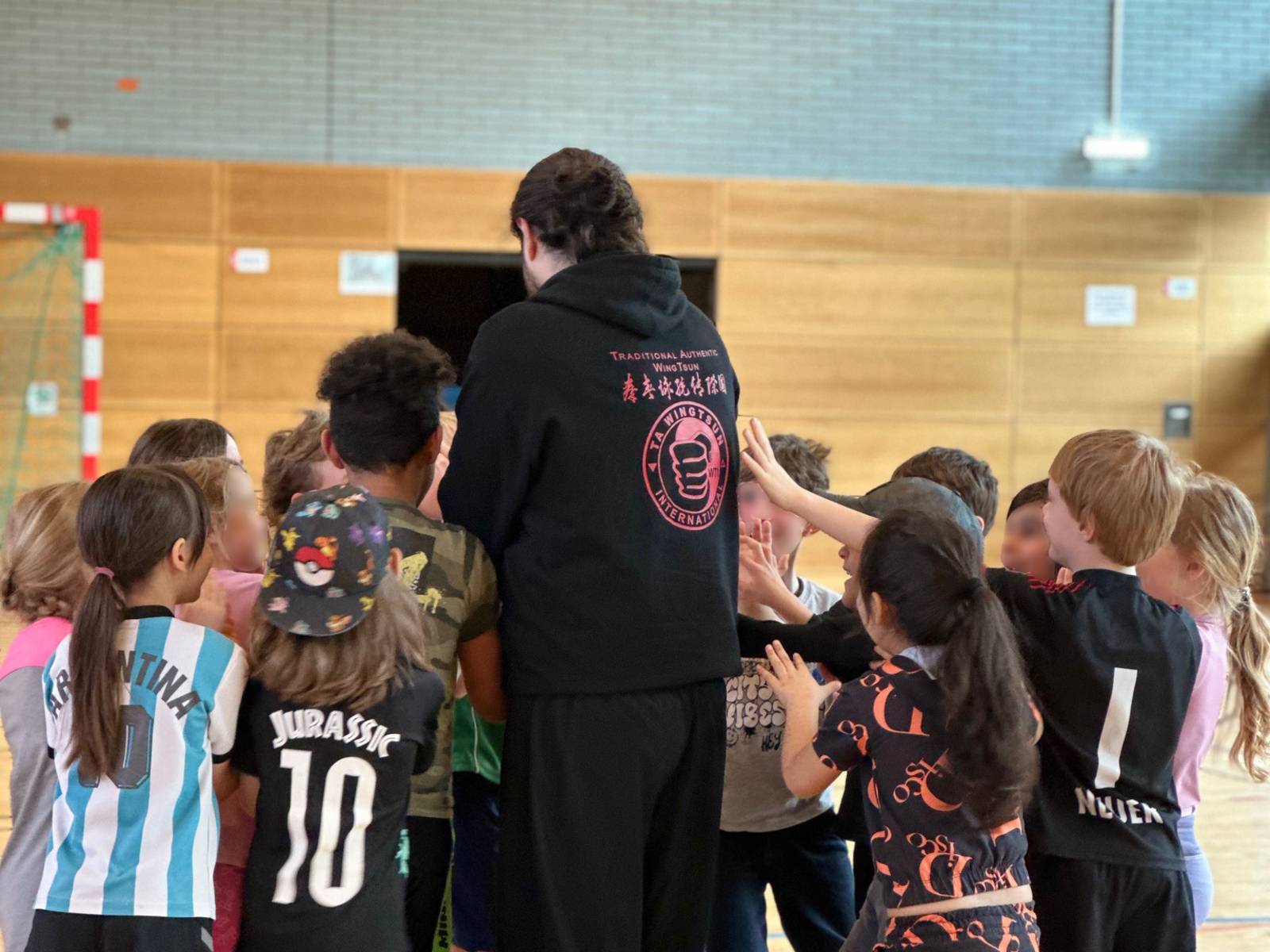 Eine Gruppe von Kindern und ein erwachsener Trainer stehen in einer Turnhalle in einem Kreis und strecken sich die Hände entgegen.