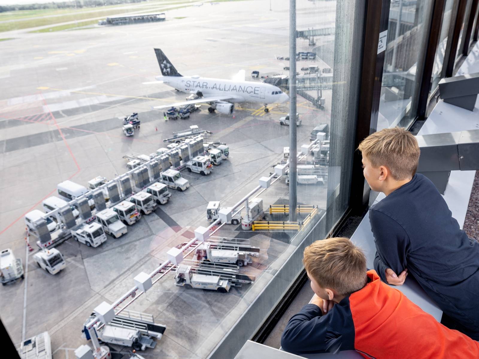 Zwei Kinder blicken von oben auf das Rollfeld eines Flughafens herab. Auf dem Rollfeld stehen neben einem Flugzeug zahlreiche andere Fahrzeuge.