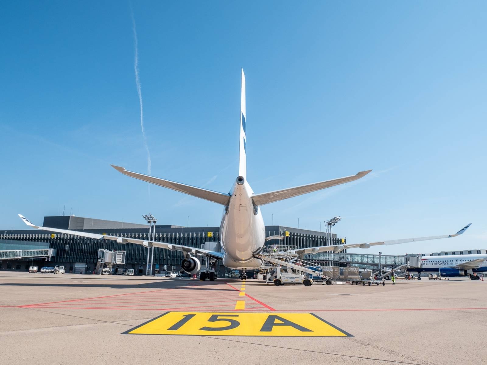 Ein auf einem Rollfeld stehendes Flugzeug ist von hinten zu sehen. Es steht vor einem Flughafengebäude. Drumherum stehen weitere Fahrzeuge und auch ein weiteres Flugzeug ist teilweise am rechten Rand zu sehen.
