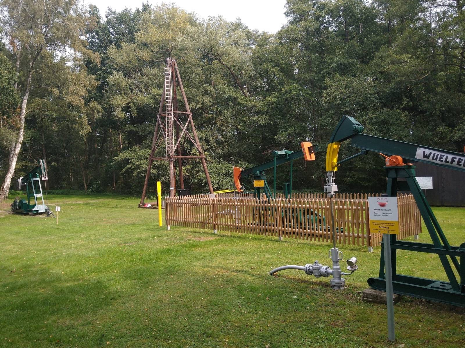 Eine alte Ölförderanlage mit Bohrturm und Pumpen auf einer Rasenfläche umgeben von Bäumen.