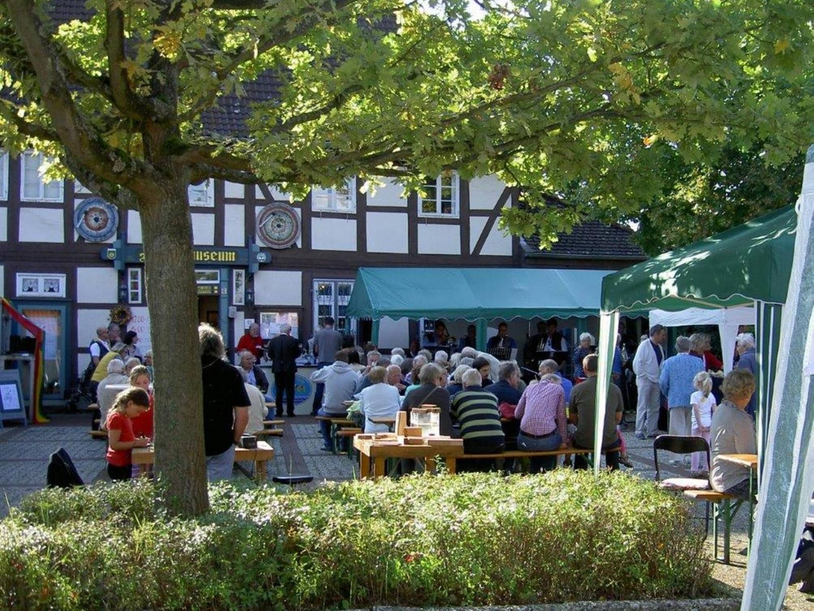 Zahlreiche Menschen sitzen auf Bierzeltgarnituren. Im Hintergrund das Heimatmuseum.