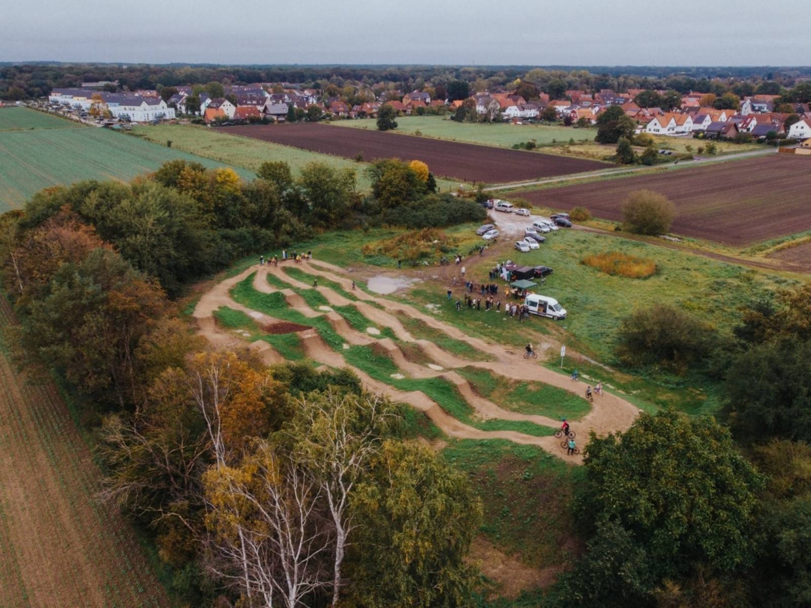 Der Dirtpark in Dedensen aus der Vogelperspektive. Auf der hügeligen Piste fahren mehrere Personen Fahrrad. Auf der Wiese neben der Piste stehen zahlreiche Menschen, Autos und ein paar Pavillons.