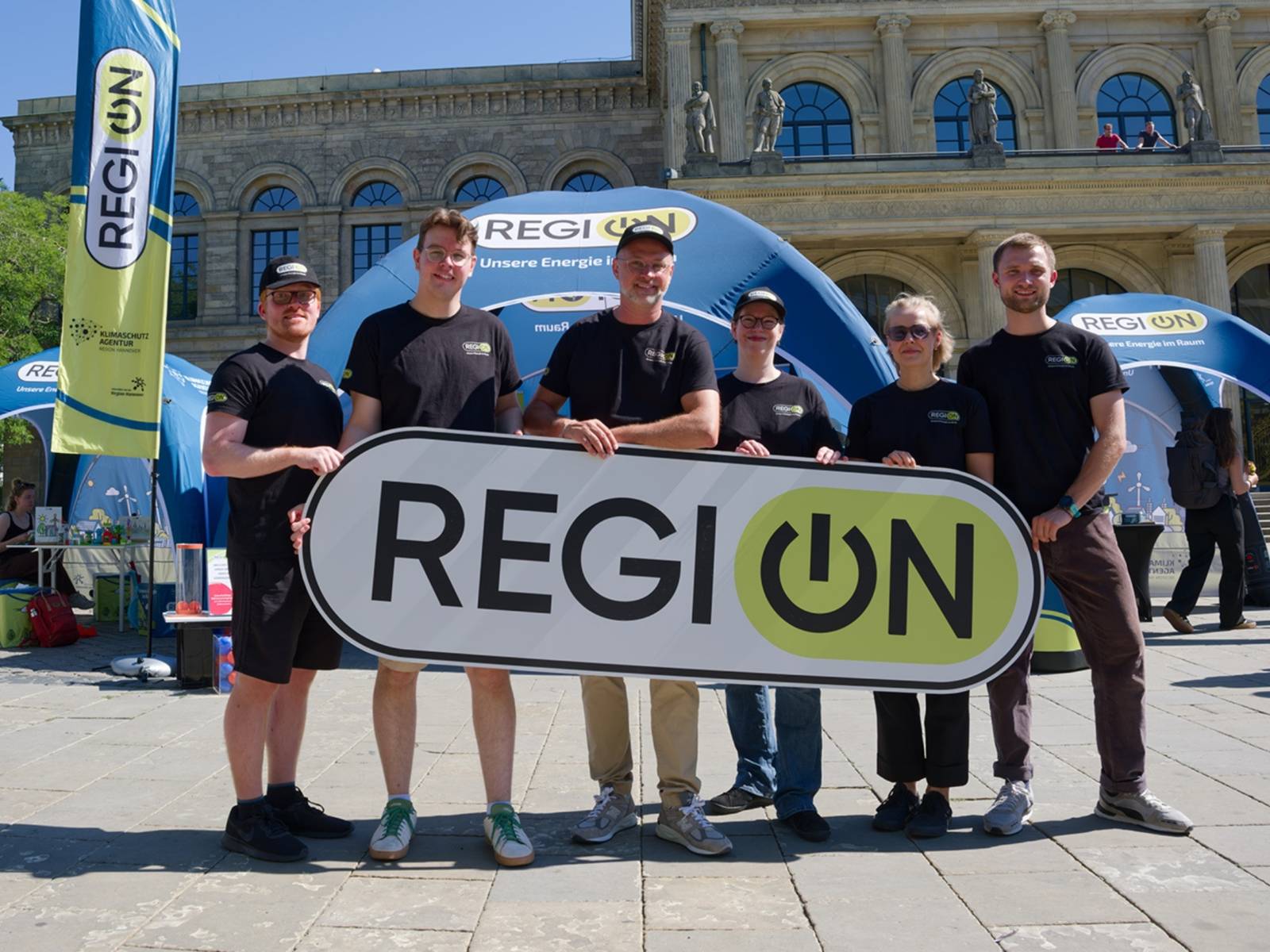 Sechs Menschen halten ein Schild mit der Aufschrift "RegiON" in den Händen, im Hintergrund sieht man Stände vor der Oper in Hannovers Innenstadt.