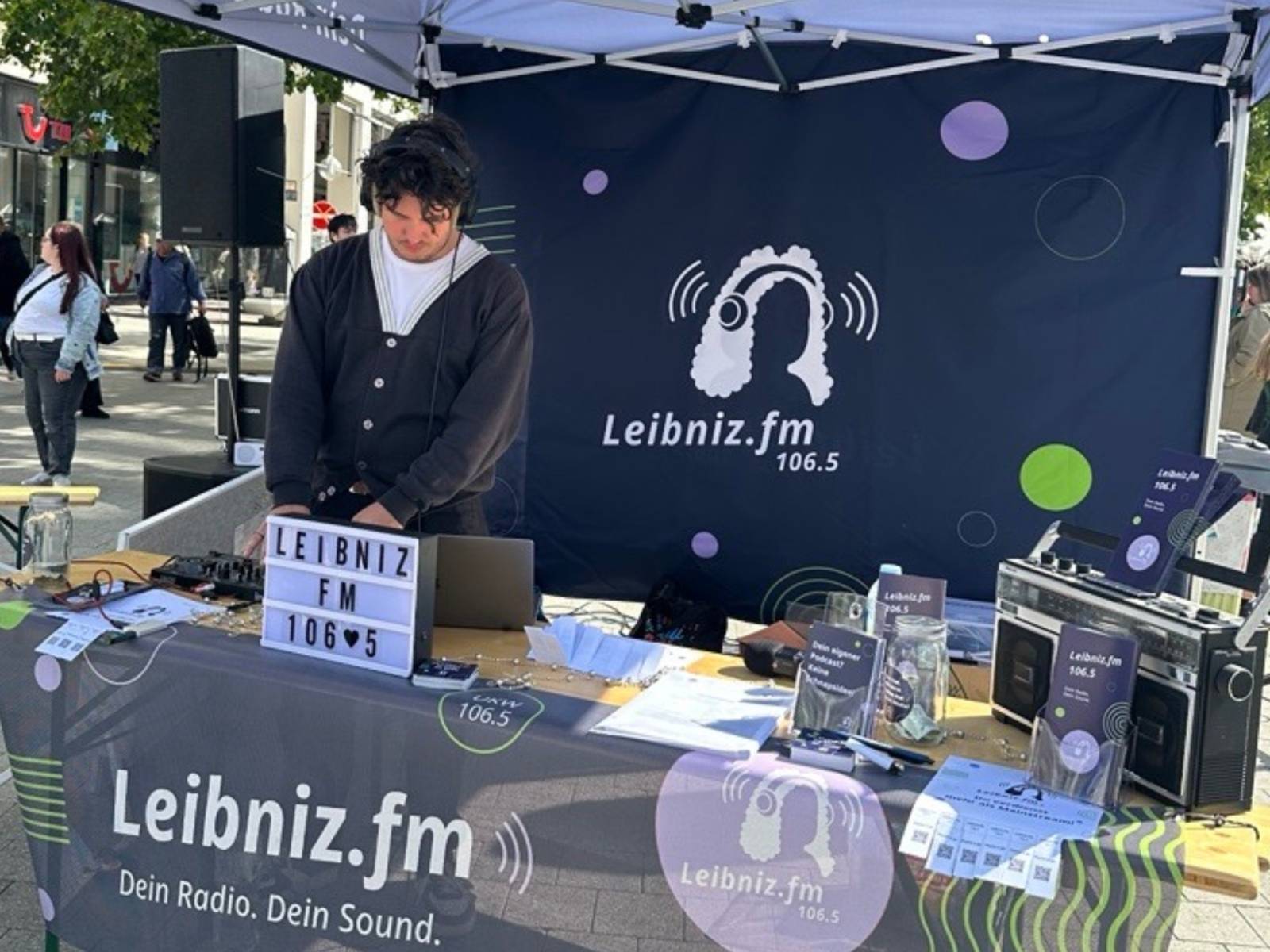 Ein Mann steht in einem Pavillon, hat Kopfhörer auf und schaut nach unten auf einen Computer mit DJ-Pult.