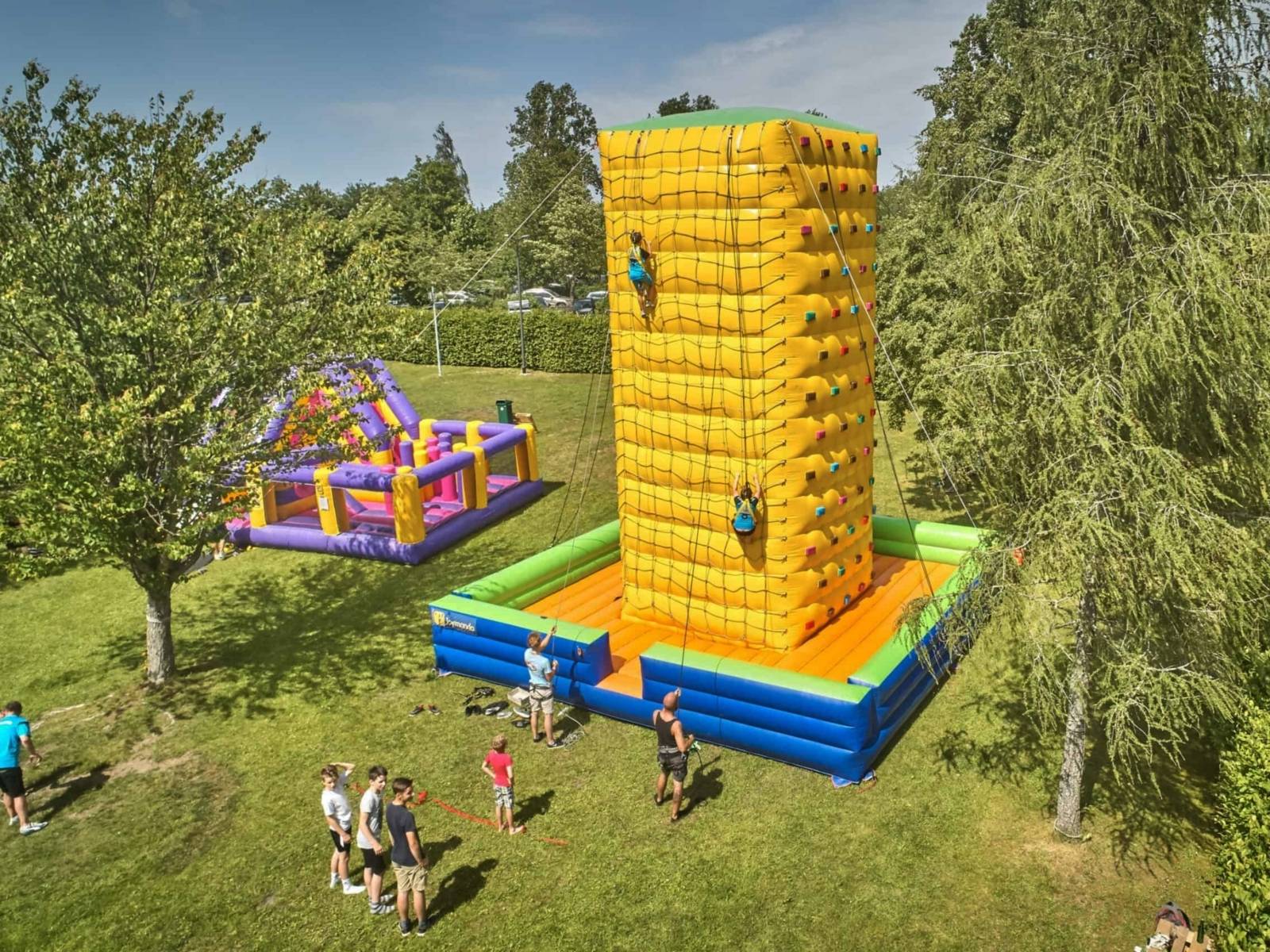Kinder und Jugendliche klettern einen bunten Kletterturm hinauf und werden dabei jeweils von einer Person mit einem Seil abgesichert. Der Kletterturm befindet sich in einem Park.