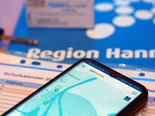 Auf einem Smartphone ist die Katwarn-App geöffnet. Das Smartphone liegt auf einem Schulkalender, drumherum sind ein Autoschlüssel, eine GVH Monatsfahrkarte und das Logo der Region Hannover.