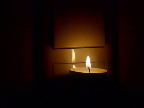 Ein Teelicht steht auf dem massiven Stecker einer LED-Lampe vor dem Lichtschalter einer Deckenbeleuchtung. Nur die Flamme bringt etwas Licht, drumherum ist es dunkel.