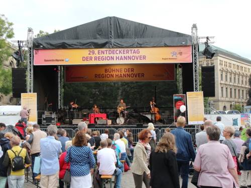 Musizierende spielen auf einer Bühne, auf einem Banner steht "29. Entdeckertag der Region Hannover. Bühne der Region Hannover". Davor stehen Menschen und folgen dem Bühnenprogramm.