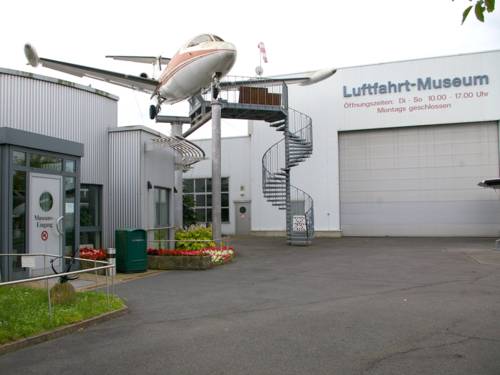 Links ist eine Glastür, rechts eine große Halle mit einem Rolltor. Dort steht der Schriftzug "Luftfahrt-Museum" und die Öffnungszeiten des Museums. Ein Flugzeug wird von Metallstreben in der Luft gehalten, eine Wendeltreppe führt zum Flugzeug.