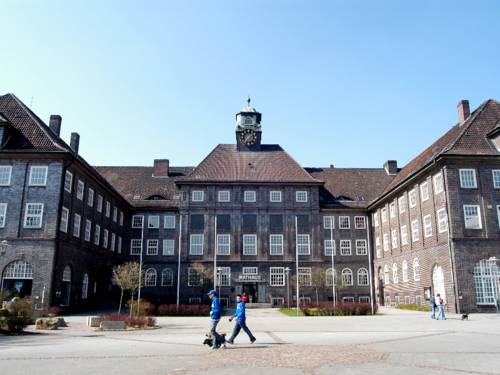 Massiver Klinkerbau mit zwei Seitenflügeln und Turmuhr vor blauem Himmel.