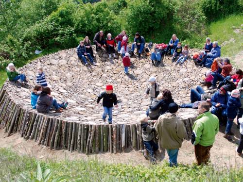 Abgesägte Baumstämme bilden eine große Schale, in der viele Menschen Platz haben. Erwachsene sitzen auf dem erhöhten Rand, Kinder klettern aus der Mitte auf den Rand.