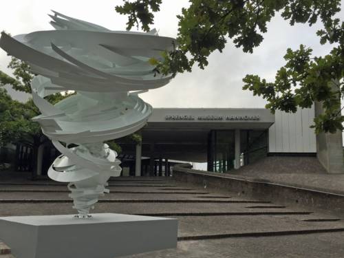Weiße Stahlskulptur mit spiralförmigen Elementen vor einem Gebäude.