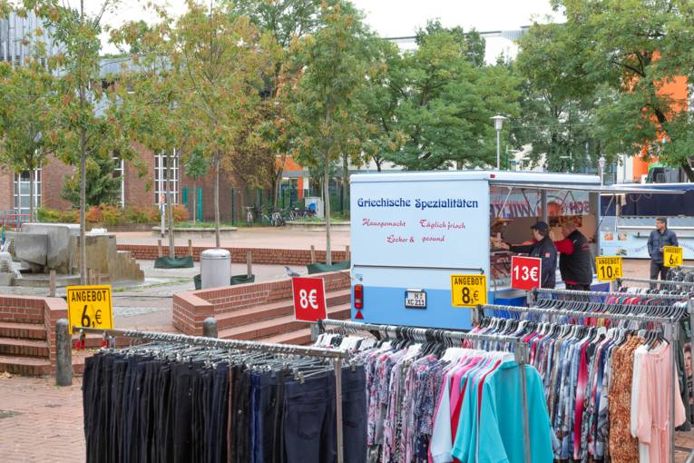 Auf einem Wochenmarktstand werden Kleidungsstücke angeboten; im Hintergrund ein Wagen mit griechischer Feinkost.