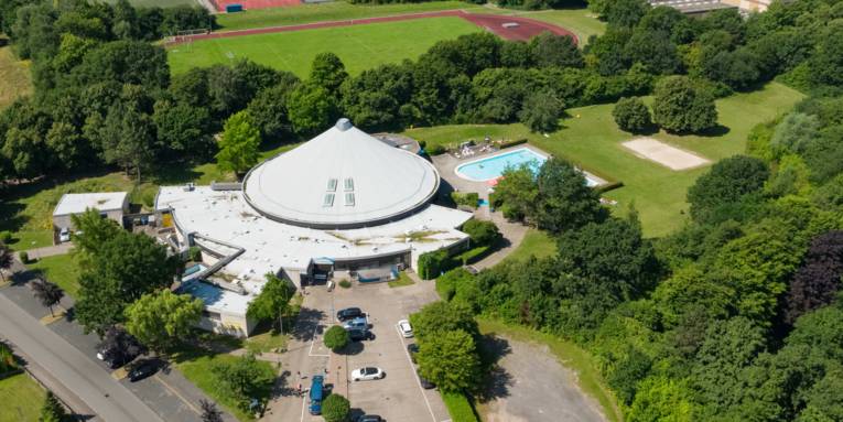 Luftbild eines Schwimmbades mit Schwimmhalle und Freibad sowie angrenzendem Sportplatz