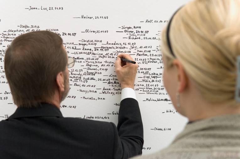 Ein Mann und eine Frau stehen vor einem White Board, der Mann schreibt etwas mit einem schwarzen Stift darauf.