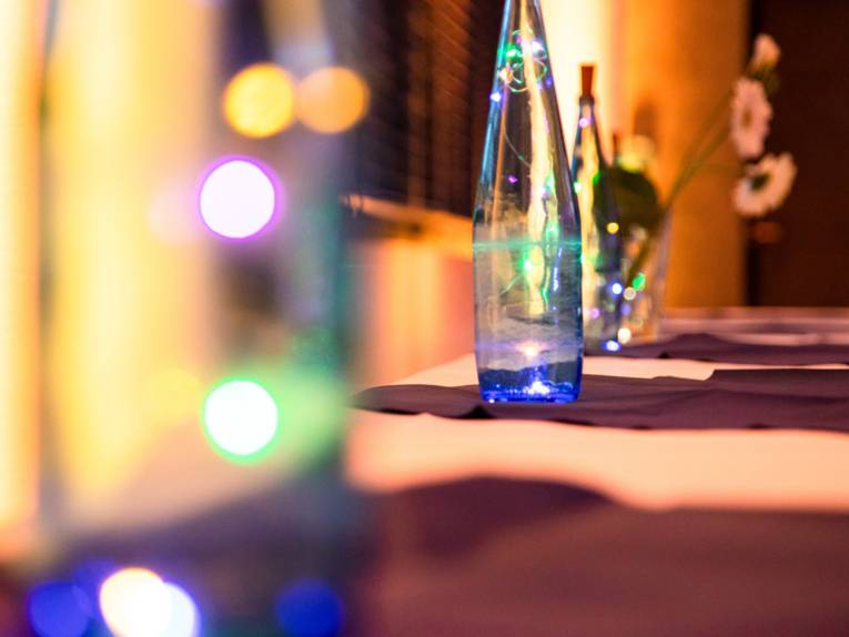 Glasflaschen und andere Gefäße stehen als Deko auf einem Tisch. Kleine LED-Lichterketten leuchten im inneren der Glasgefäße und setzen verschiedene Farbpunkte.