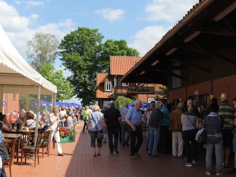 Zahlreiche Menschen befinden sich auf dem Innenhof eines Bauernhofes. Einige von ihnen sitzen an Tischen unter Pavillons und Sonnenschirmen. Andere stehen am Tresen unter dem großen Vordach einer Scheune.