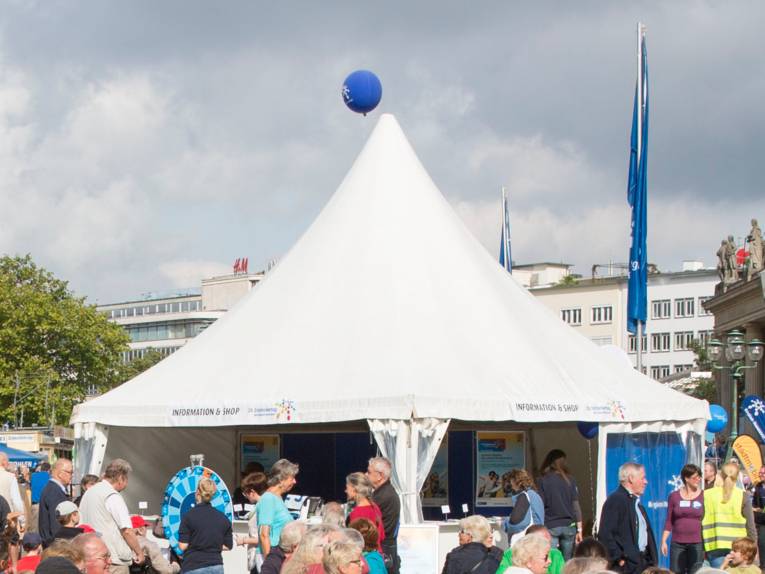 Weißes Pavillonzelt mit zwei offenen und zwei geschlossenen Seiten. Drumherum und drinnen sind Menschen.