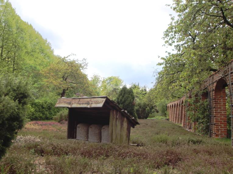 Eine kleine Bretterbude mit schrägem Dach und drei Holtwänden schützt drei Bienenkörbe vor Wind und Regen, drumherum wächst Heide.