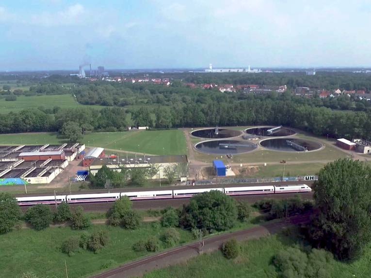 Blick auf die verschiedenen Klärbecken des Klärwerks Herrenhausen. Davor fährt ein ICE vorbei.