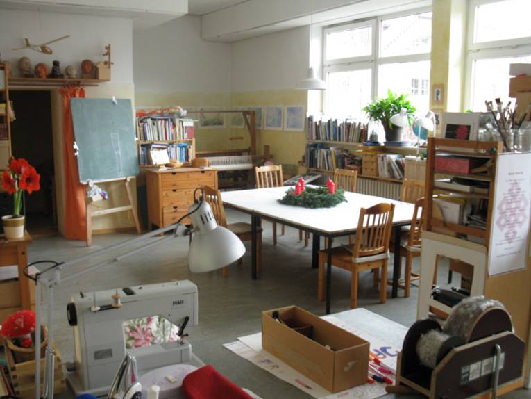 Mitten in einem Raum steht ein großer Tisch mit sechs Holzstühlen dran. In dem Raum befinden sich eine Tafel, Regale mit Bücher, eine Nähmaschine und viele Bastel- und Hobbyutensilien.