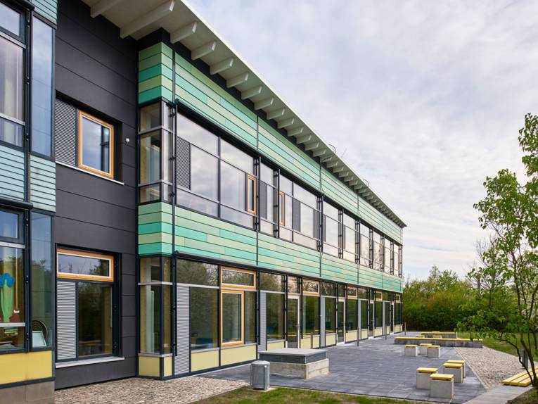 Die Außenansicht der Grundschule An der Feldbuschwende zeigt einen zweistöckigen Flachbau mit großen Fensterfronten und grüner Verkleidung, davor einen Pausenbereich mit Sitzelementen.