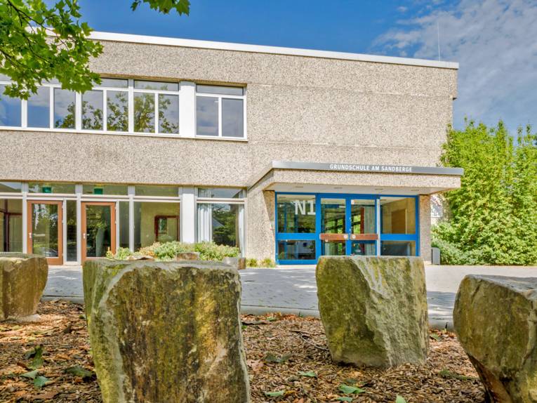 Der Eingang zur Grundschule am Sandberge mit blauen Türen in einem zweistöckigen Gebäude mit großen Fensterfronten und Waschbetonverkleidung.