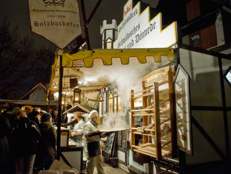 Ein Mann in Bäckerkleidung holt vor zahlreichen Zuschauern ein dampfendes Blech aus einem Holzbackofen.