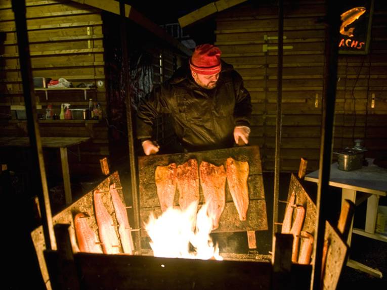 Ein kräftiger Mann mit roter Mütze trägt eine Holzkonstruktion, auf der Lachsfilets zum Räuchern hängen.