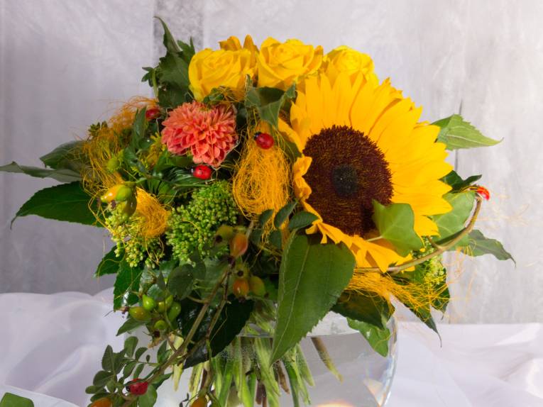 Ein Blumenstrauß in Gelbtönen mit roten Akzenten: Sonnenblume, Dahlie, Rosen, Hagebutten und Sedum mit Blattgrün werden von roten Marienkäfern geziert.