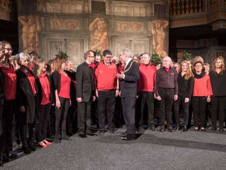 Chor auf einer Bühne in einer Galerie. Davor viele Menschen.