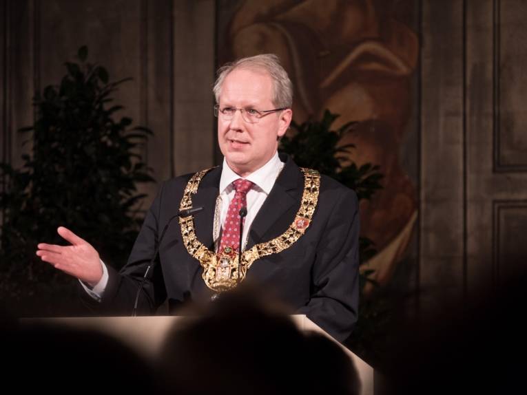 Mann mit einer Amtskette auf einer Bühne. Er spricht zu vielen Menschen.