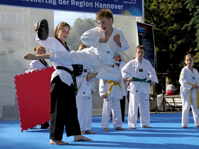 Ein Junge im weißen Kampfsportanzug fliegt durch die Luft und tritt mit dem Fuß gegen einen gepolsterten Handschuh, den eine Trainerin festhält. Im Hintergrund sind weitere Kinder im Kampfsportanzug vor dem Banner der Sportaktionsfläche zu sehen.