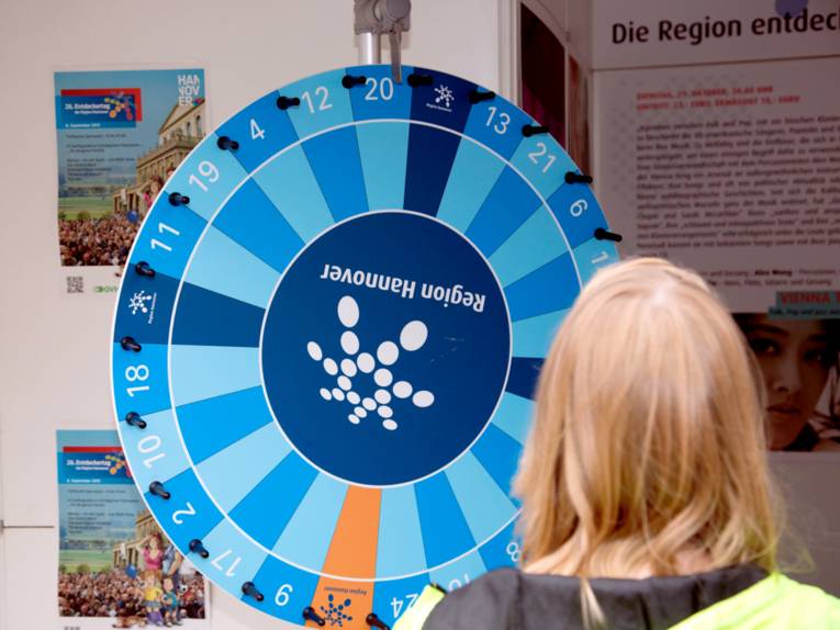 Ein Mädchen steht vor einem Glücksrad. Die Felder des Glücksrades sind überwiegend blau, in der Mitte ist das Punktelogo der Region Hannover.
