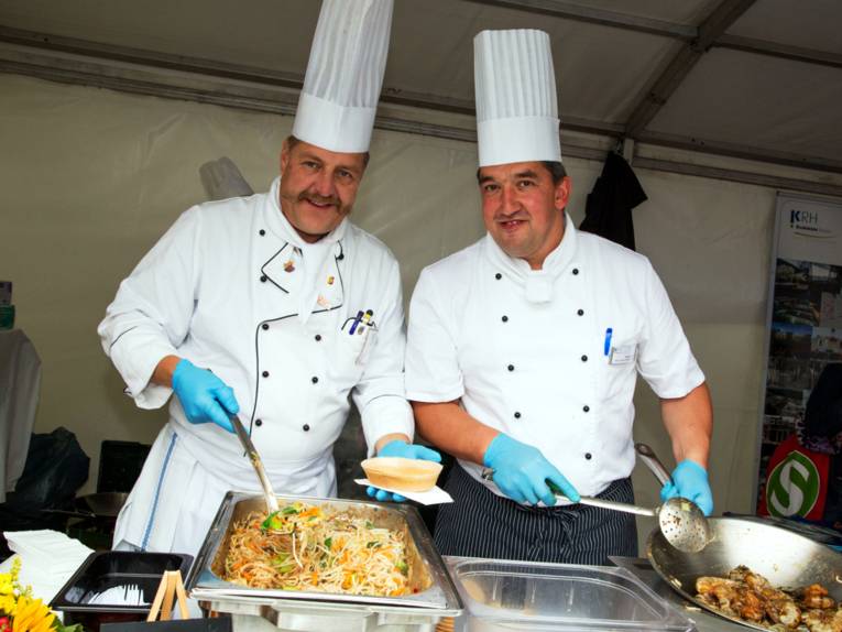 Zwei Männer tragen weiße Kochkleidung und hohe Kochmützen, sie bieten ein Nudelgericht und Scampis aus einem großen Metallbehälter und einer Pfanne an.