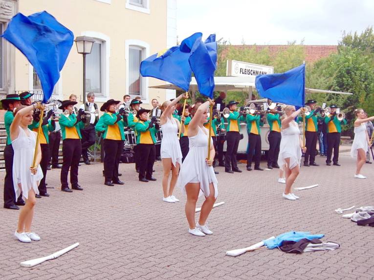 Mädchen der Colorguards tragen weiße Kleider und schwenken blaue Fahnen, dahinter spielt ein Fanfarenzug.