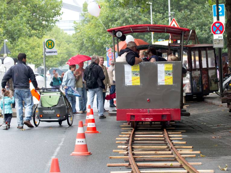Schienen liegen auf der Georgstraße, eine kleine Lokomotive fährt darauf und befördert Menschen. Auf der Straße sind Fußgänger unterwegs.