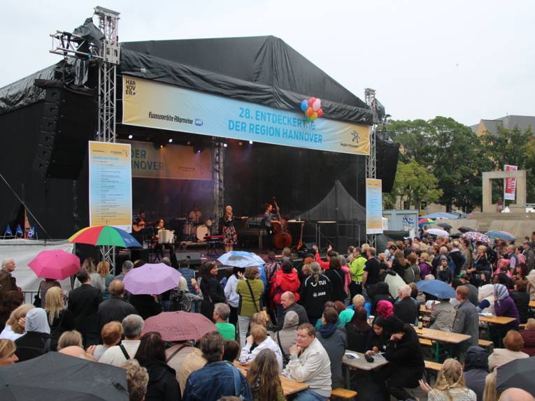 Entdeckerinnen und Entdecker folgen dem Bühnenprogramm, vorsorglich sind einzelne Regenschirme gegen erste Tropfen aufgespannt.