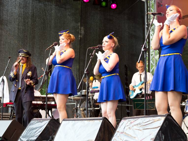 Drei Frauen tragen blaue Kleider, die an die Uniform einer Flugbegleiterin erinnern und singen in Mikrofone. Daneben steht ein Mann, der die Fantasieuniform eines Flugkapitäns trägt und ebenfalls in ein Mikrofon singt.