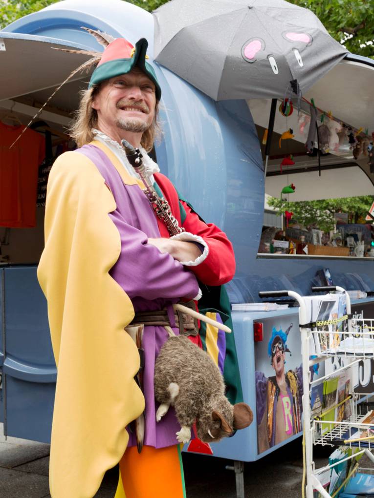 Ein Mann trägt ein buntes Kostüm, an seinem Gürtel baumelt eine Plüschratte und vor der Brust hält er eine Flöte. Hinter ihm ist ein Marktwagen mit Souvenirs der Stadt Hameln.