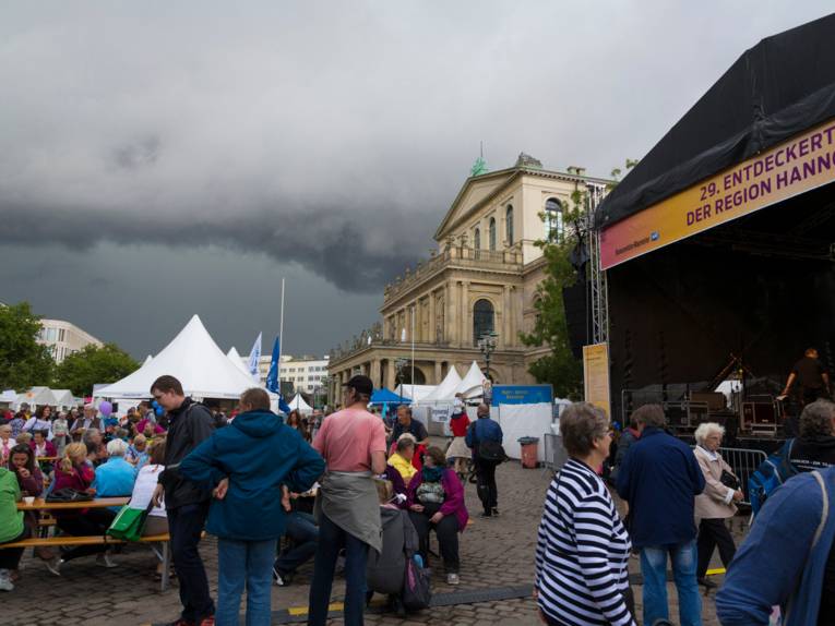 Eine große Bühne und Zelte sind auf dem Opernplatz in Hannover, im Hintergrund ist das Operngebäude. Am Himmel ziehen sich dunkle Wolken zusammen. Menschen gehen über den Platz, stehen oder sitzen an Festzeltgarnituren.