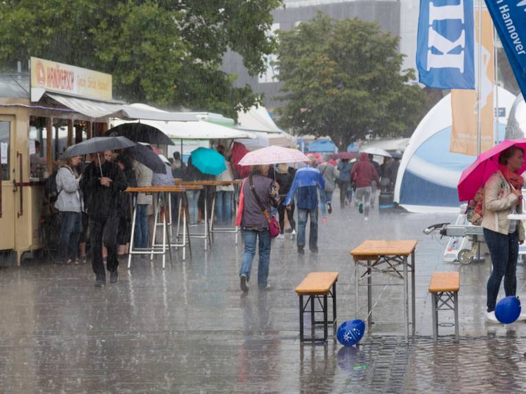Auf dem Opernplatz in Hannover stehen Zelte und Infostände. Es regnet stark und es bilden sich Pfützen auf dem Pflaster. Menschen gehen unter aufgespannten Regenschirmen durch den Regen.
