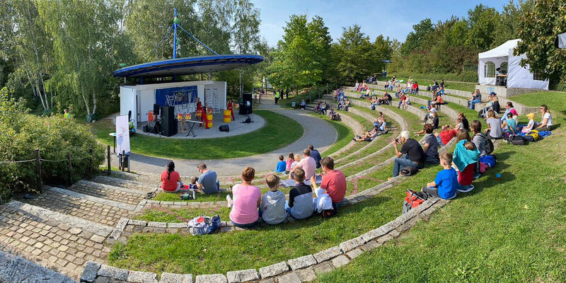 Menschen sitzen mit Abstand in einem Rondell und verfolgen das Programm auf einer Bühne. Menschen sitzen mit Abstand in einem Rondell und verfolgen das Programm auf einer Bühne.