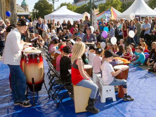 Kinder und ein Erwachsener trommeln vor dem Opernhaus. Der Boden ist für Artistinnen und Artisten des Kinder- und Jugendcircus Bunttropfen mit Turnmatten ausgelegt. Kinder und ein Erwachsener trommeln vor dem Opernhaus. Der Boden ist für Artistinnen und Artisten des Kinder- und Jugendcircus Bunttropfen mit Turnmatten ausgelegt.