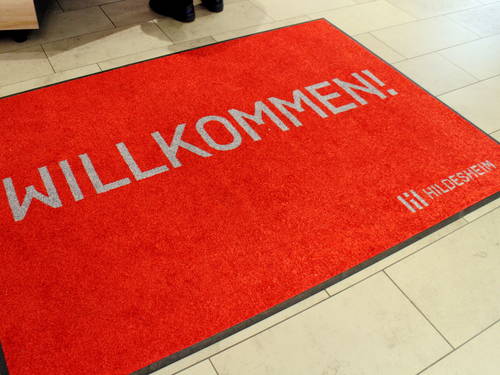 Eine rote Fußmatte liegt auf dem gefliesten Fußboden. Darauf steht in grauer Schrift: "Willkommen! Hildesheim". Eine rote Fußmatte liegt auf dem gefliesten Fußboden. Darauf steht in grauer Schrift: "Willkommen! Hildesheim".