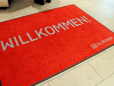 Eine rote Fußmatte liegt auf dem gefliesten Fußboden. Darauf steht in grauer Schrift: "Willkommen! Hildesheim". Eine rote Fußmatte liegt auf dem gefliesten Fußboden. Darauf steht in grauer Schrift: "Willkommen! Hildesheim".