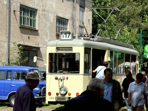 An einer Haltestelle steigen einige Fahrgäste in eine historische Straßenbahn, weitere Personen schlendern auf dem Bahnsteig. An einer Haltestelle steigen einige Fahrgäste in eine historische Straßenbahn, weitere Personen schlendern auf dem Bahnsteig.