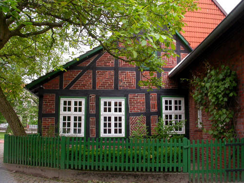 Hinter einem Holzzaun mit grünen Latten steht kleines Fachwerkhaus. Die Räume zwischen den dunklen Holzbalken sind mit roten Mauersteinen ausgefüllt. Hinter einem Holzzaun mit grünen Latten steht kleines Fachwerkhaus. Die Räume zwischen den dunklen Holzbalken sind mit roten Mauersteinen ausgefüllt.