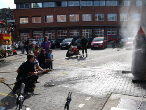 Kinder und Jugendliche spritzen mit D-Strahlrohren in die Fenster eines Brandhauses, links steht ein Löschfahrzeug und im Hintergrund ist das Gebäude der Feuer- und Rettungswache 1 zu sehen. Kinder und Jugendliche spritzen mit D-Strahlrohren in die Fenster eines Brandhauses, links steht ein Löschfahrzeug und im Hintergrund ist das Gebäude der Feuer- und Rettungswache 1 zu sehen.